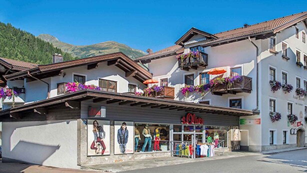 A hotel with a storefront displaying clothing, balconies with flower boxes, and a mountainous backdrop. - Holiday apartment with infrared (Rauris)
