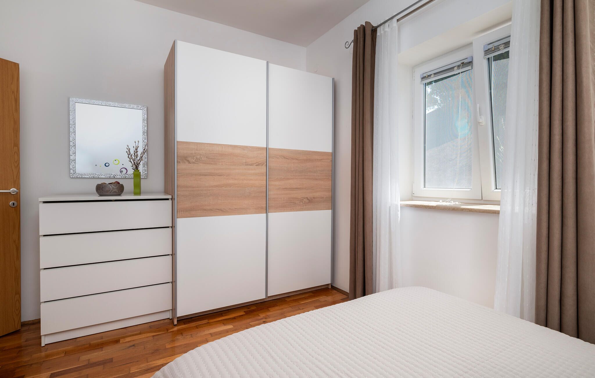 A bedroom with a bed, a wardrobe with a mirror, a dresser, and a window with curtains.