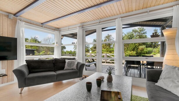 Een ruime woonkamer met een grijze bank, een bijzettafeltje met decoratieve voorwerpen, en een televisie. Grote ramen bieden uitzicht op een houten terras en groen.
