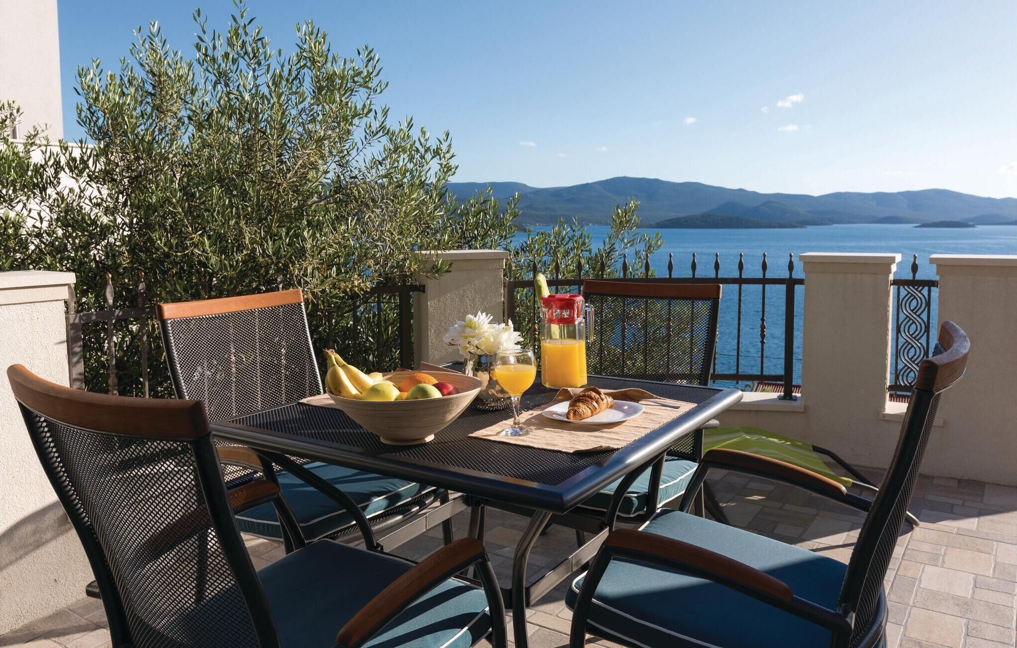 Ein Tisch mit Früchten, einem Croissant und einem Glas Orangensaft auf einem Balkon mit Blick auf das Meer und die Berge.