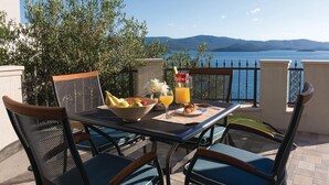 A table set with fruits, a croissant, and a glass of orange juice on a balcony with a view of the sea and mountains.