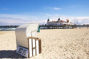 Ein Strandstuhl mit der Aufschrift "80 35" auf einem sandigen Strand, im Hintergrund ein Steg und Gebäude.