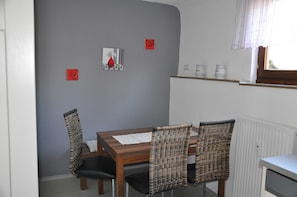 Un coin repas avec une table en bois et des chaises en osier, un mur gris orné d’une rose rouge et une fenêtre avec des rideaux blancs.