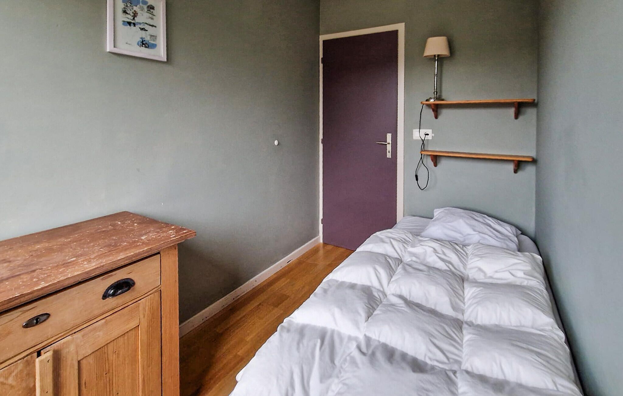 A bedroom with a bed, wooden dresser, door, and wall-mounted lamp.