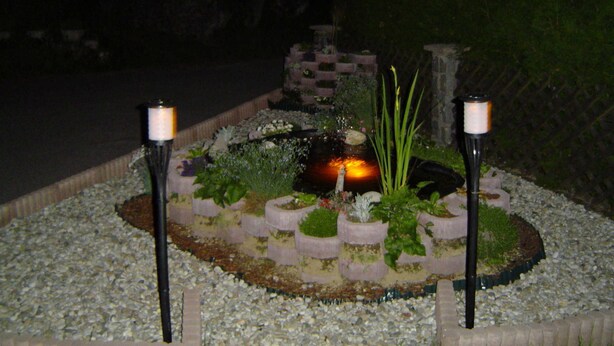 Een tuin met een vijver, omringd door planten in potten en twee lichtjes die op kaarsen lijken.