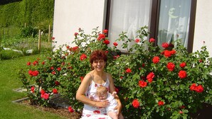 Een vrouw houdt een baby vast voor een huis met een tuin vol rode rozen.