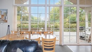 A dining area with a large table and chairs, a leather couch, and a view of greenery through large windows.