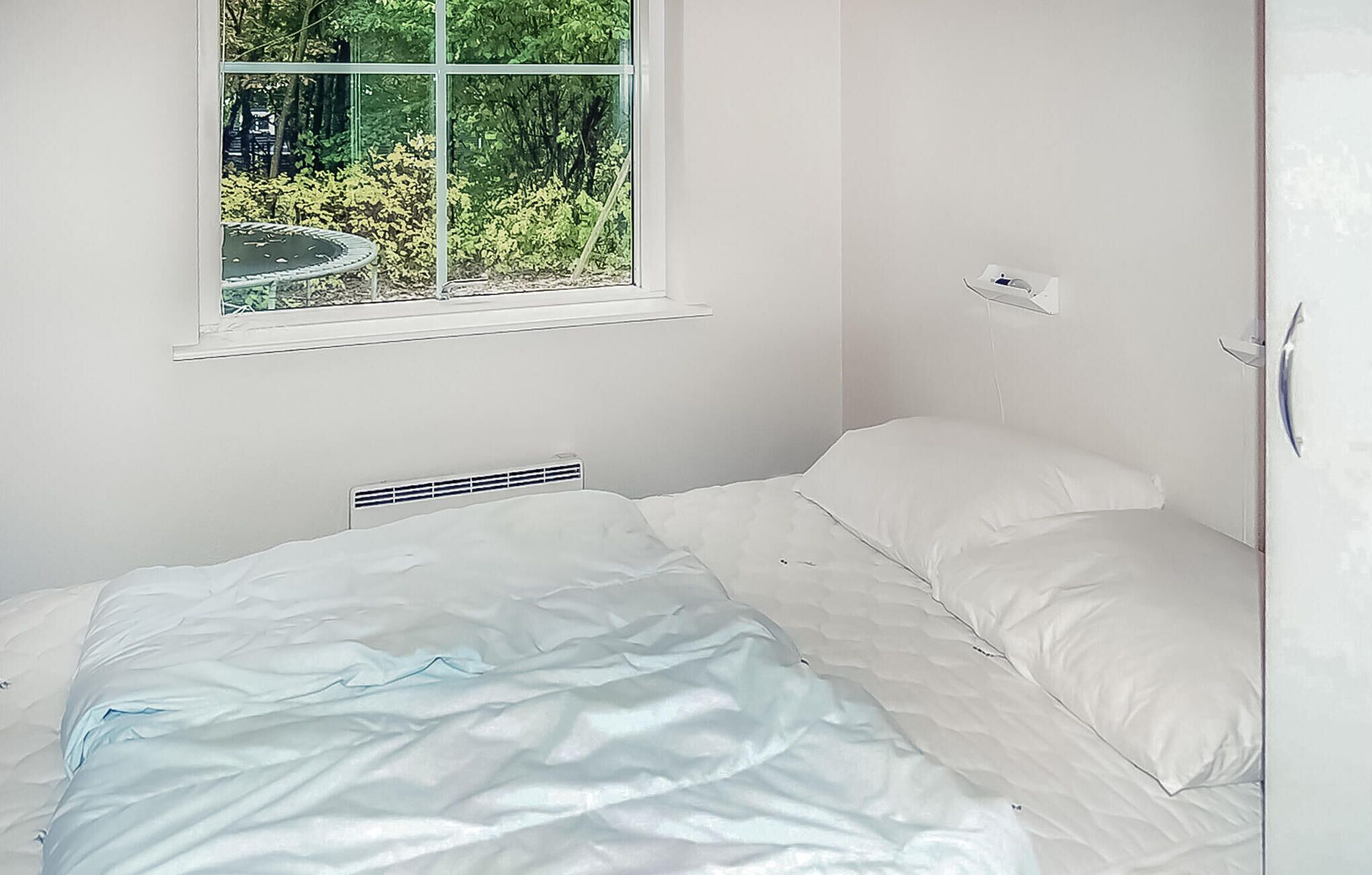 Ein kleiner, weißer Raum mit einem Einzelbett, einem Fenster mit Blick ins Grüne und einem Thermostat an der Wand.