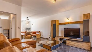 Un salon moderne avec un canapé en cuir, une table basse en verre, un meuble de rangement en bois intégrant un téléviseur à écran plat, et un coin repas avec une table et des chaises en bois.