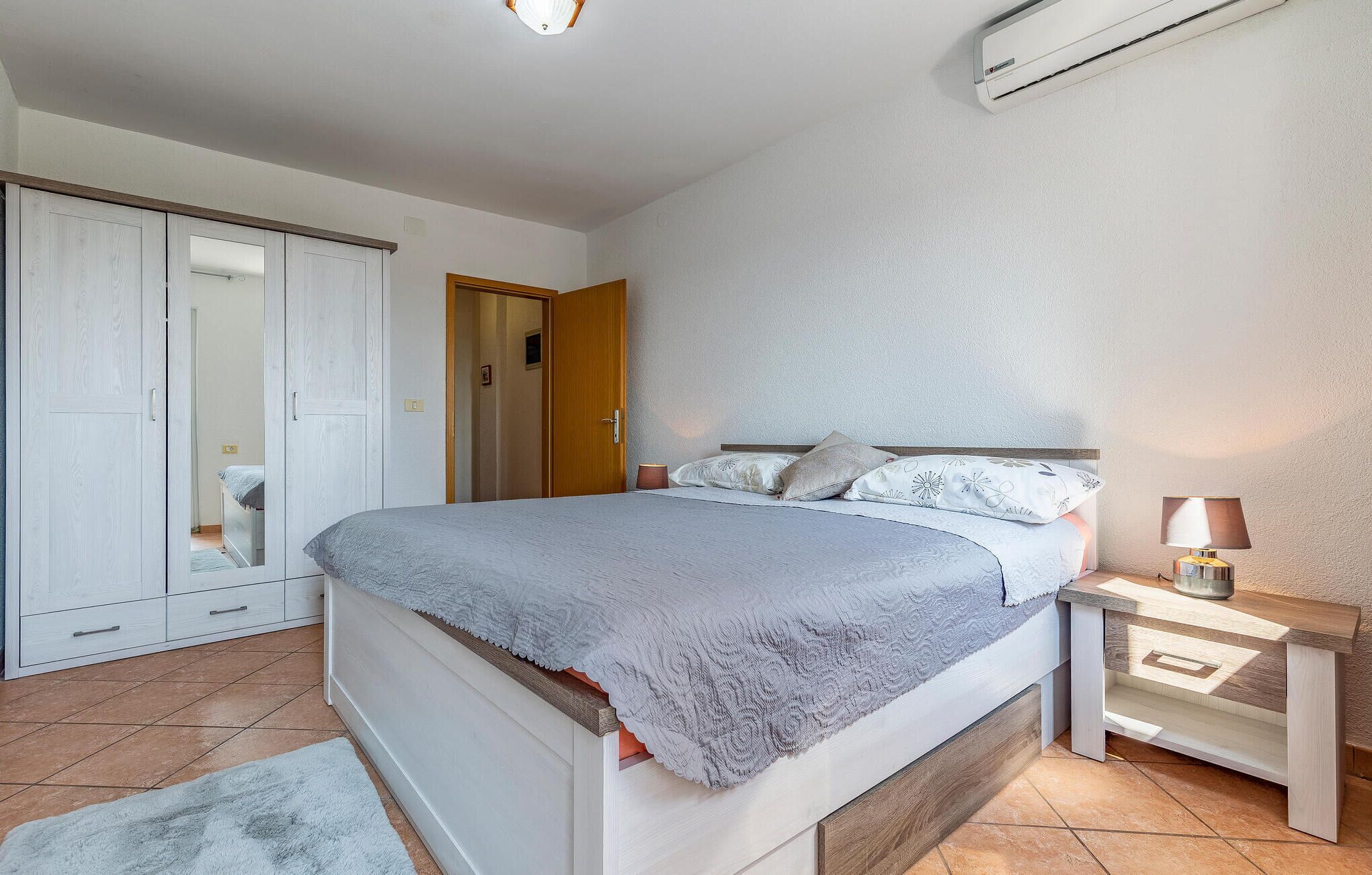 A bedroom with a bed, bedside table, lamp, wardrobe, and an air conditioning unit.