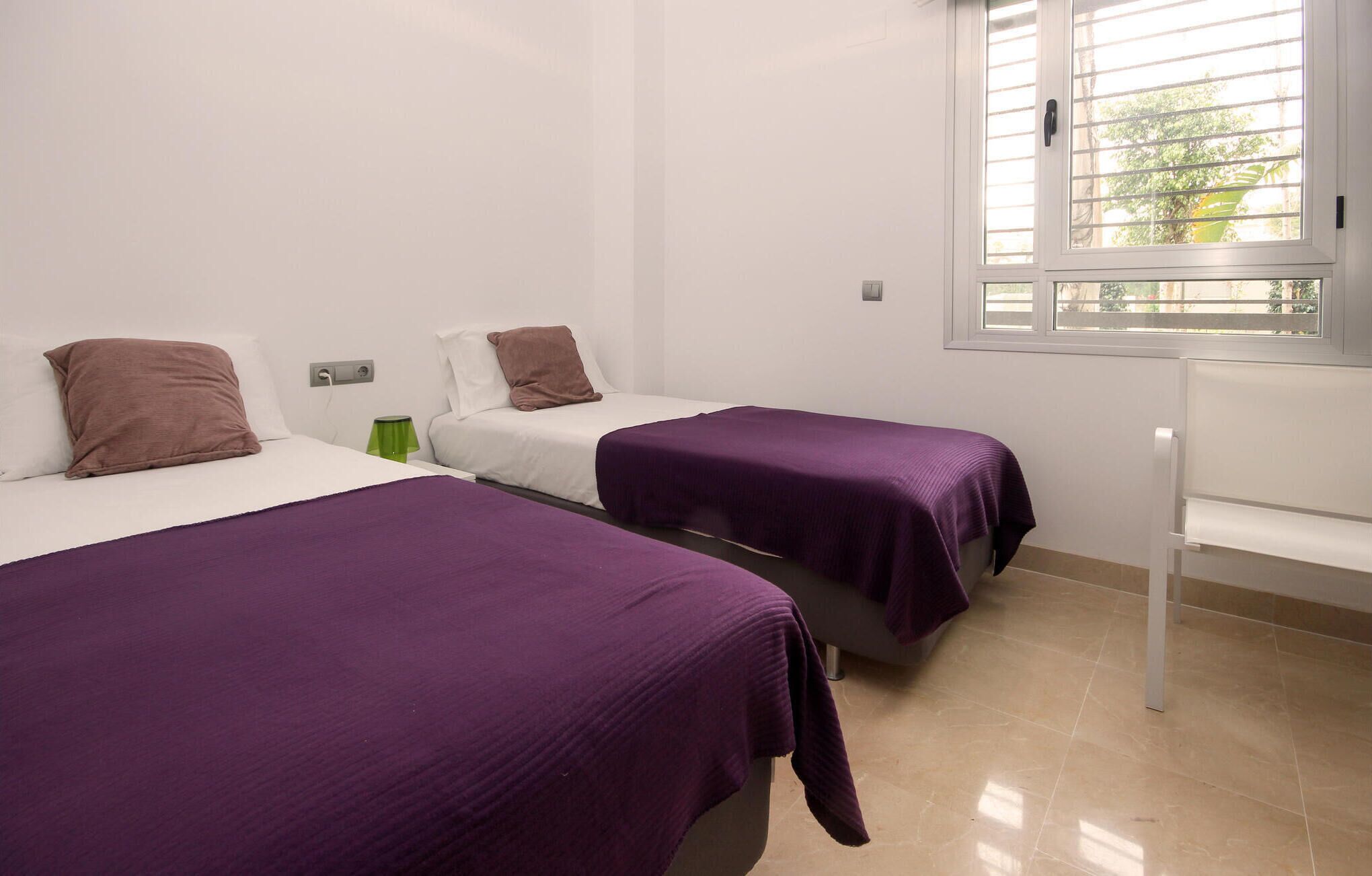 A room with two beds, each with a purple bedspread and brown pillows, a green lamp, and a window with blinds.