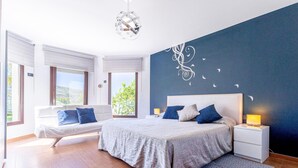 Quarto moderno com uma cama grande, mesas de cabeceira, um sofá e vista para o verde lá fora.