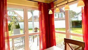 Ein Schlafzimmer mit einem Bett, einem Stuhl und einem Tisch. Große Fenster mit Vorhängen bieten Blick ins Freie.