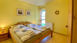 Ein Doppelbett mit zwei Kissen und einer Decke, ein Nachttisch mit Lampe sowie ein Fenster mit Blick ins Freie.
