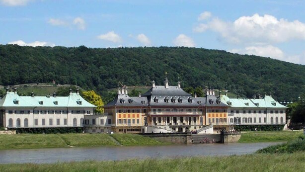 Un grand bâtiment historique, doté de plusieurs ailes et d’une cour intérieure, situé au bord d’une rivière, avec une colline boisée en arrière-plan.