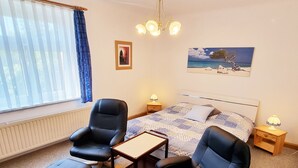 Une chambre à coucher avec un lit, deux chaises, une petite table, une table de chevet, une lampe, un tableau représentant une plage et une fenêtre avec des rideaux.