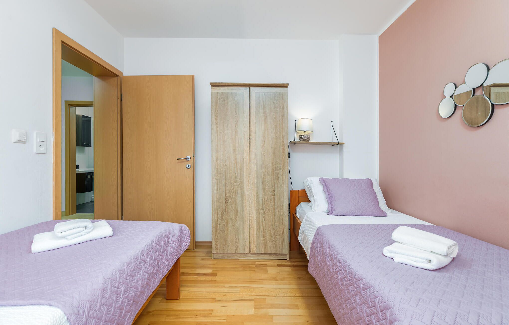 A bedroom with two single beds, a wooden wardrobe, a bedside table with a lamp, and a wall-mounted mirror.