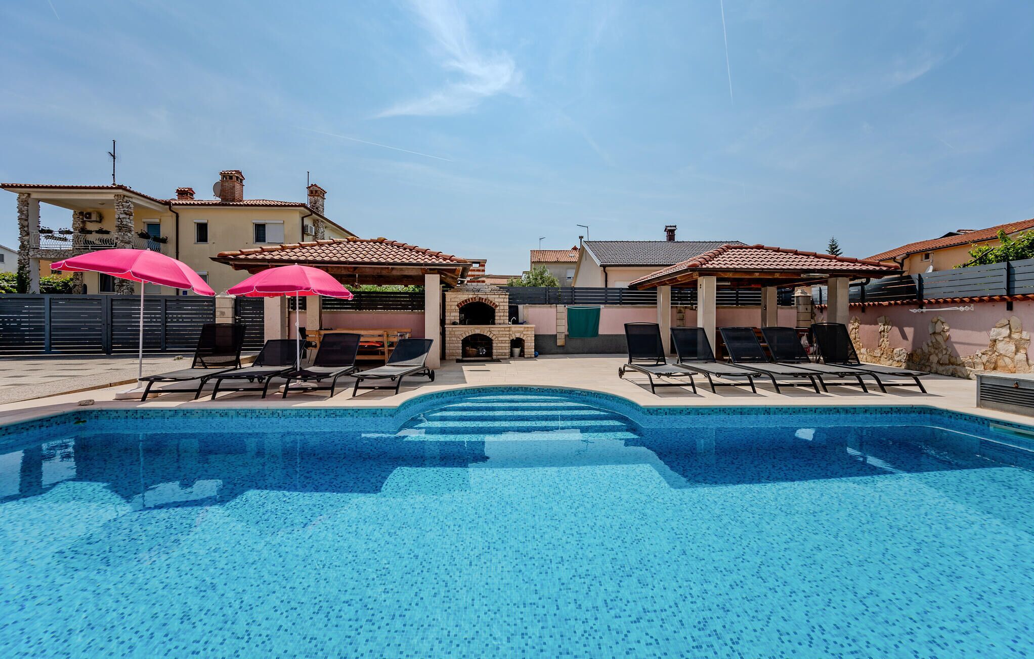 A swimming pool with lounge chairs and umbrellas, surrounded by buildings with tiled roofs.