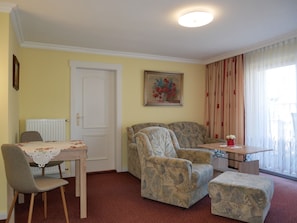 A living room with a sofa, armchair, and ottoman, a dining table with a chair, a radiator, a door, and a window with curtains.