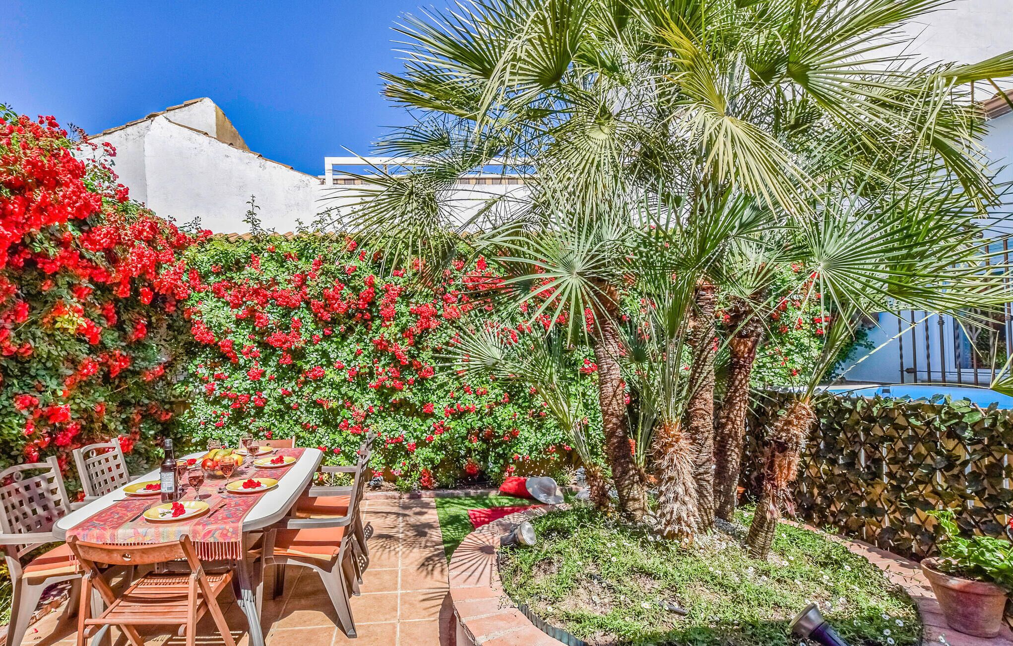 A patio with a table set for dining, surrounded by lush greenery and flowering plants.