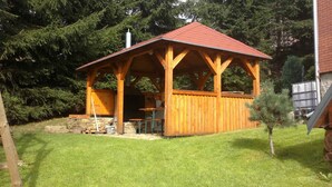 Een houten tuinhuisje met een rood dak, stenen fundering en een kleine tafel met stoelen erbij.