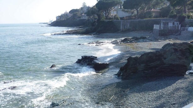 A rocky shoreline with waves crashing onto the beach, a clear sky, and buildings in the background.
