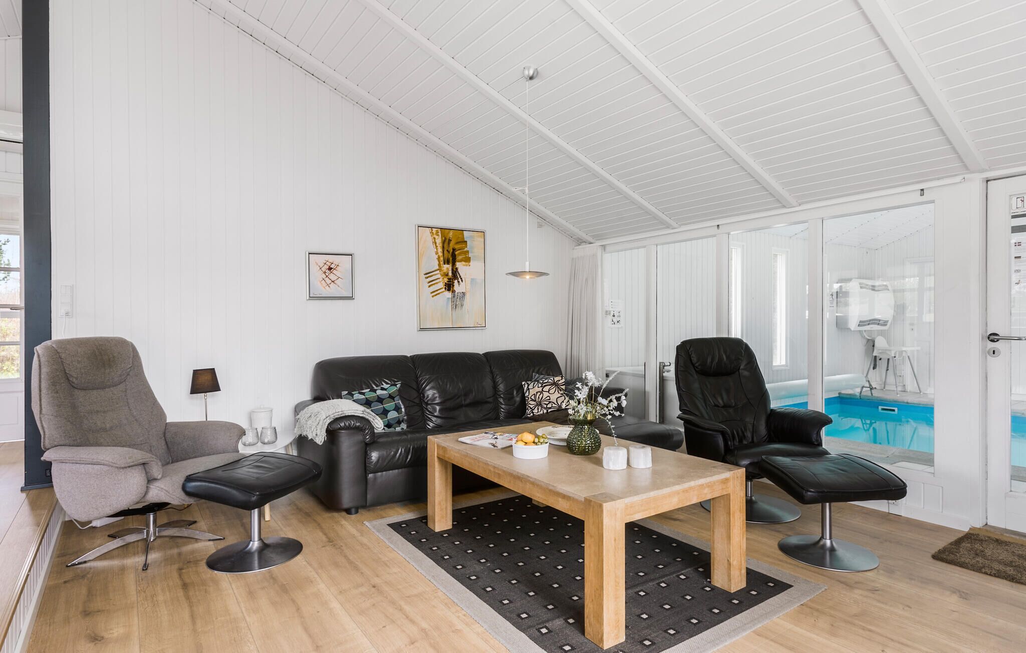 Een moderne woonkamer met een houten salontafel, twee banken, een zwarte leren fauteuil, en een zwembadje zichtbaar door een glazen deur.