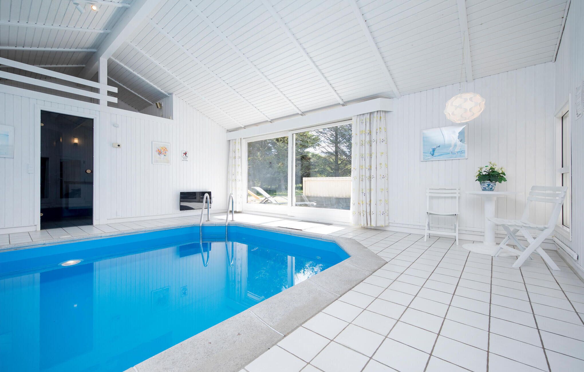 En swimmingpool med kristallklart blått vatten, omgiven av vita väggar och ett kaklat golv. Det står två vita stolar och ett bord nära poolen.