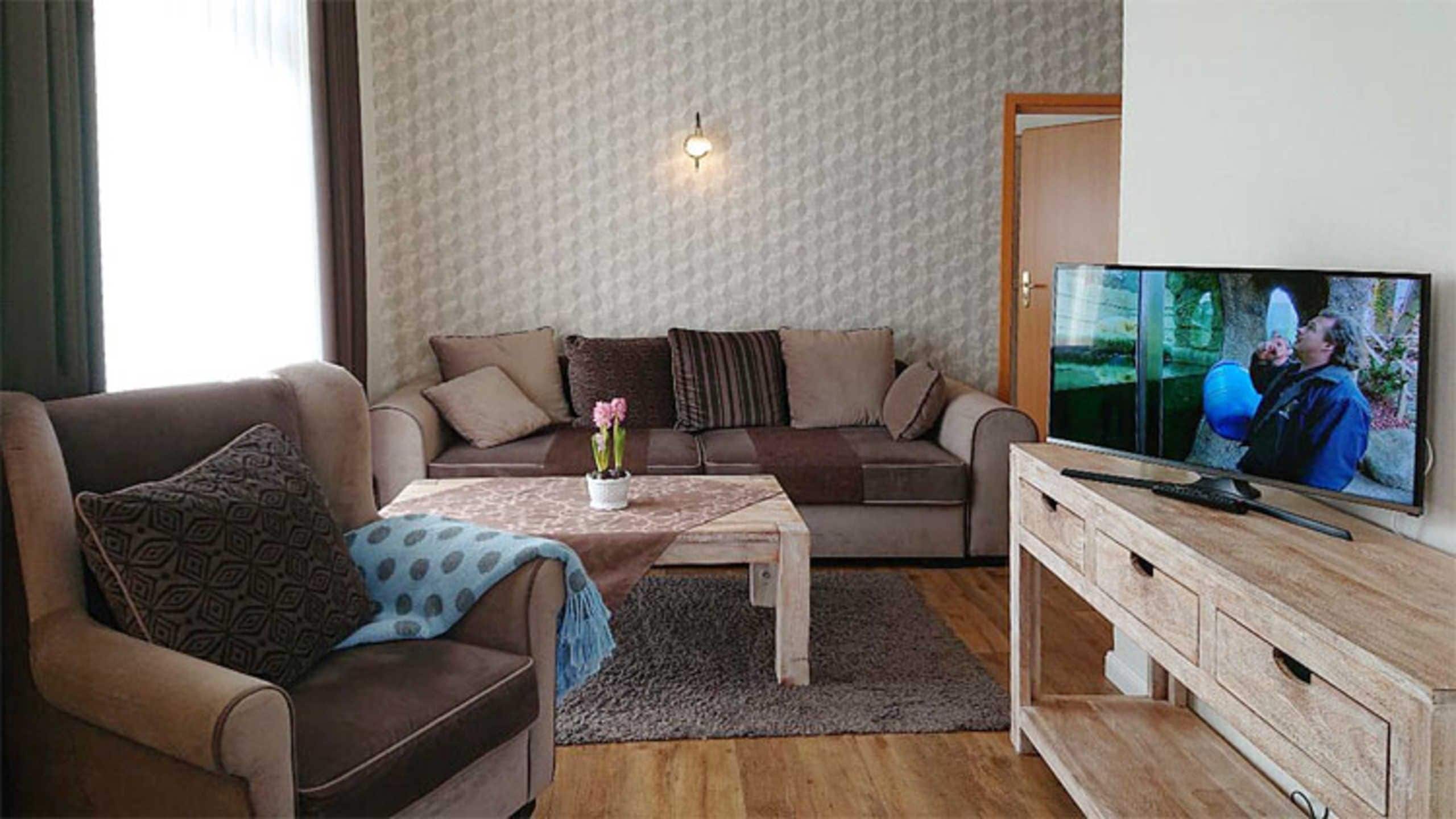 Ein Wohnzimmer mit einer Couch, einem Couchtisch mit einer Vase, einem Fernseher auf einem Holzgestell und einer Wand mit Muster.