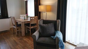 Una habitación con una mesa de comedor, sillas, un sofá con un cojín estampado, una lámpara de pie y un ventanal con cortinas.