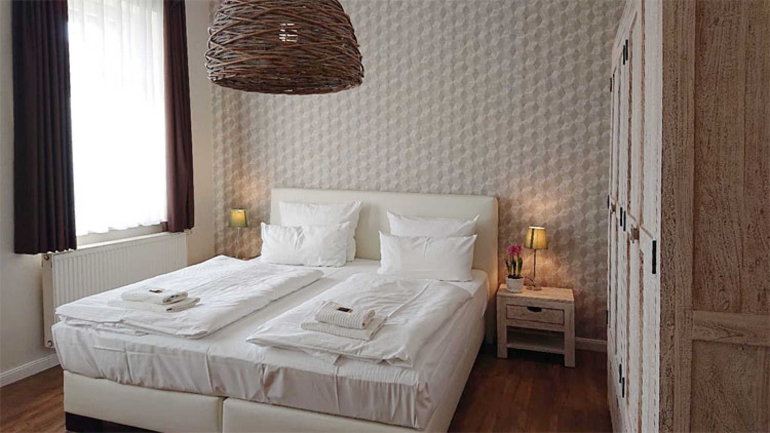 Uma cama de casal com lençóis brancos, duas mesas de cabeceira com luminárias, um lustre tecido pendurado e um papel de parede estampado.