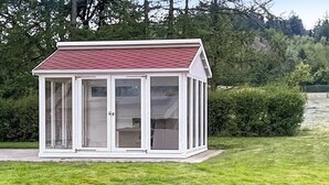 A small, white, glass-enclosed structure with a red roof, likely a greenhouse or conservatory, situated on a grassy lawn with dense trees in the background. - 3 bedroom nice home in Brovst (Brovst)