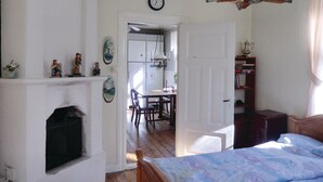 Un dormitorio con cama, chimenea, zona de comedor con mesa y sillas, y cocina visible a través de una puerta abierta.