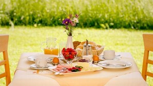 Ein Tisch im Freien mit einer Tischdecke, einer Karaffe Orangensaft, einer Schüssel Erdbeeren, Brot und einem Blumenstrauß.