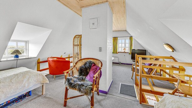 Une pièce avec un plafond en pente, un lit, une chaise rouge, une chaise en bois avec un coussin en fourrure, un téléviseur et une fenêtre avec des rideaux.
