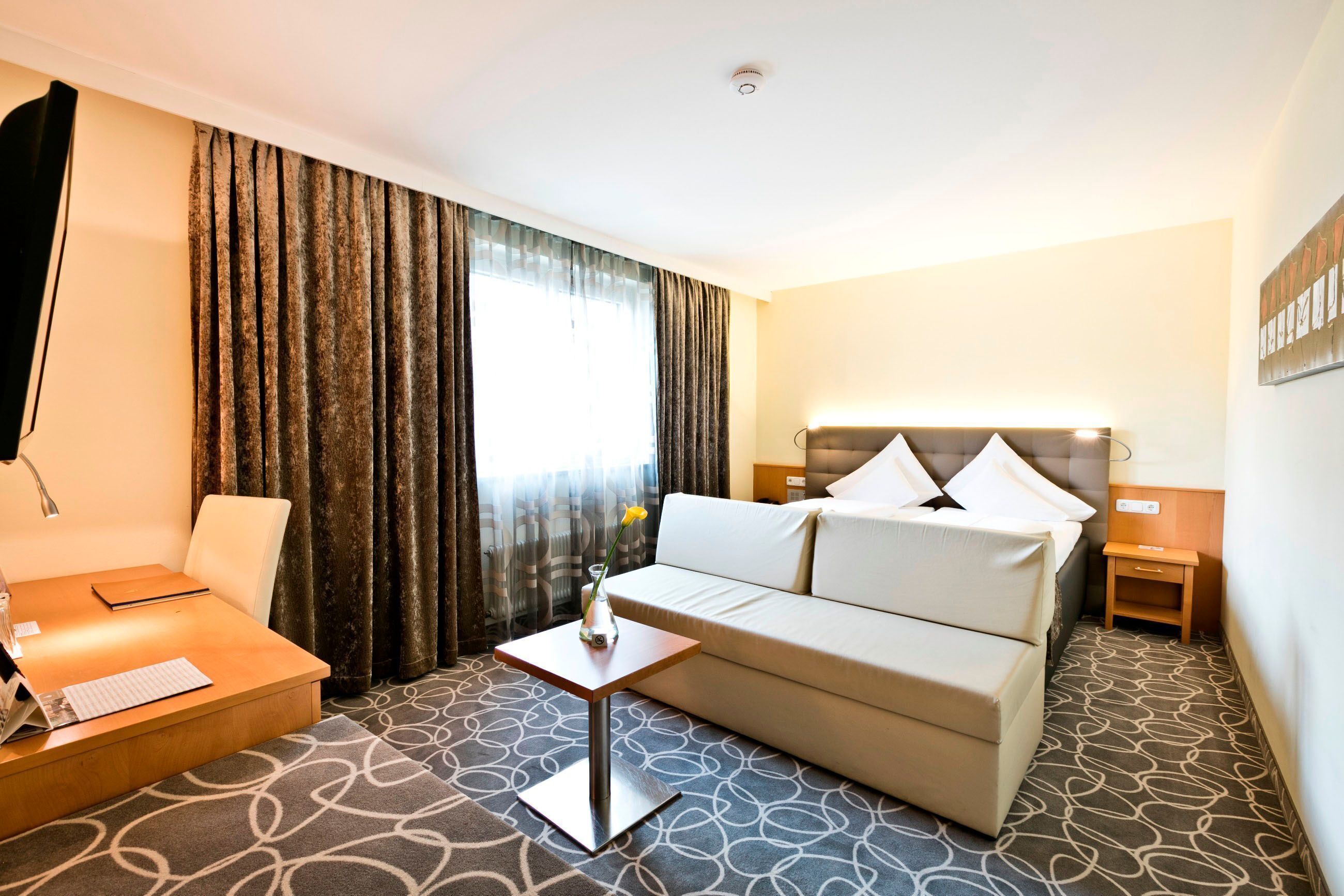 Ein Hotelzimmer mit Bett, Sofa, einem kleinen Tisch mit Vase, einer Lampe und einem Schreibtisch mit Tastatur.