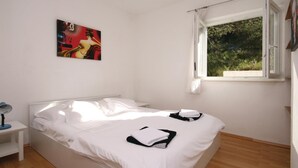Un dormitorio con cama, mesita de noche, lámpara, ventana con vistas a zonas verdes y un cuadro en la pared.