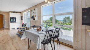 A dining area with a table set for four, a kitchen with white cabinets, and large windows offering a view of greenery and a house.