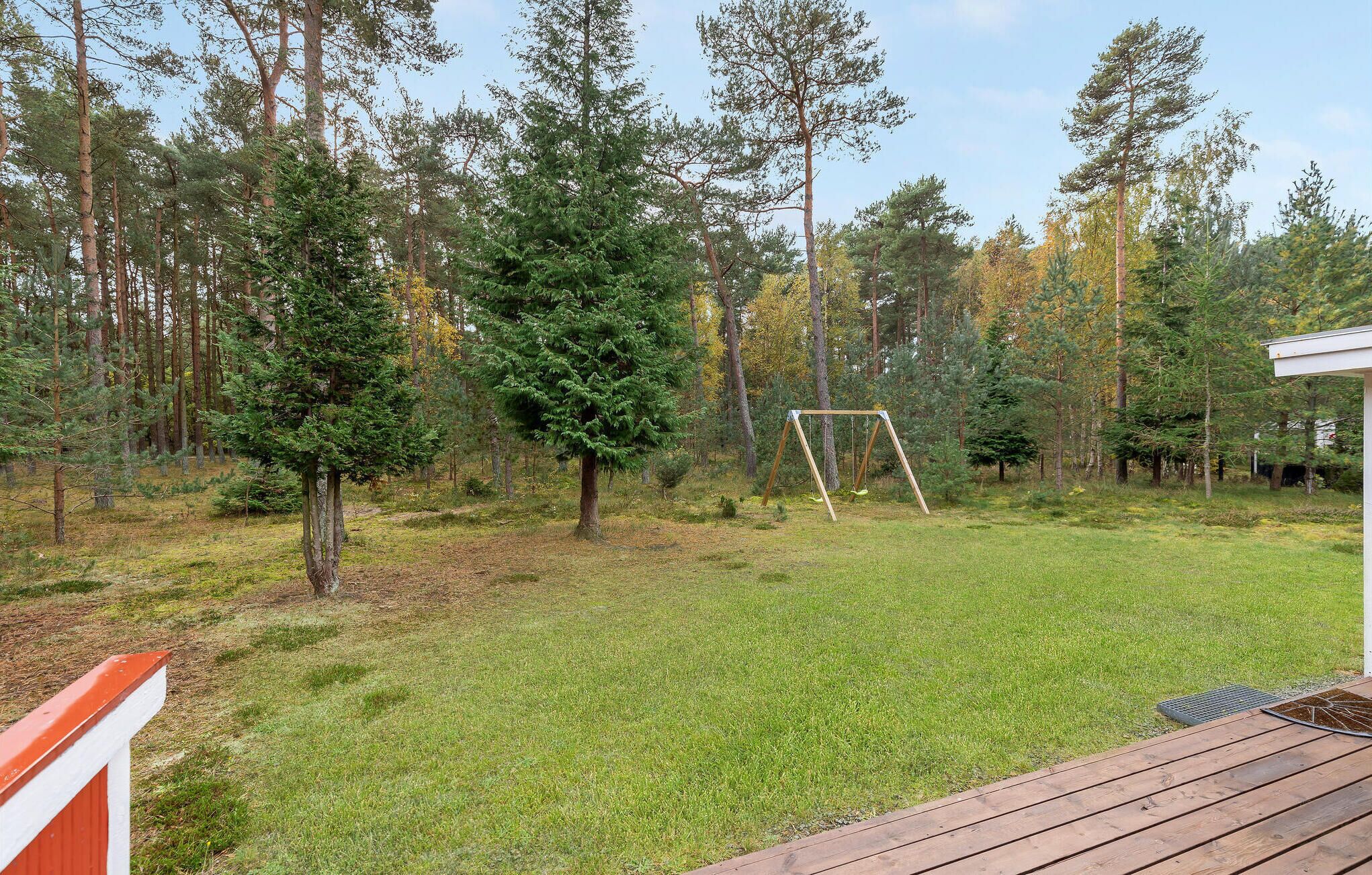 A backyard with a wooden deck, a swing set, and a grassy lawn surrounded by tall trees.