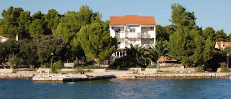 A two-story building with a red-tiled roof, surrounded by lush greenery and a waterfront.