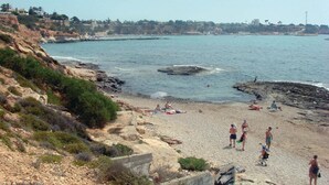 Ein Strand mit felsigem Gelände, ein paar Personen und eine entfernte Küstenlinie mit Gebäuden.