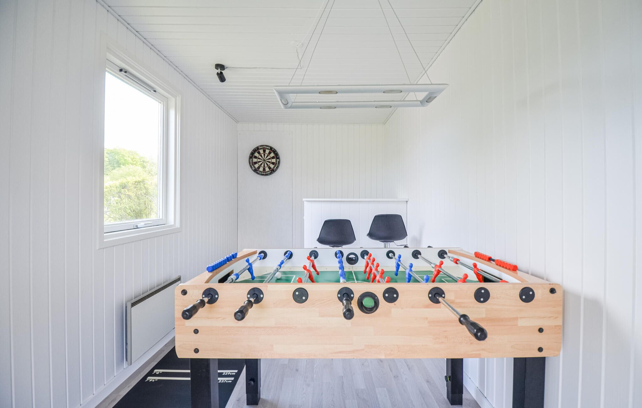 A room with a foosball table, a dartboard, and a window with a view of greenery.