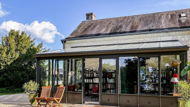 Une véranda dotée d’un toit en verre, de chaises en bois et d’étagères remplies de livres.