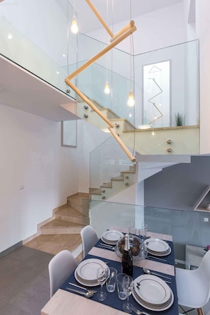 A modern dining area with a glass staircase, a wooden handrail, and a table set for six with plates, glasses, and cutlery.