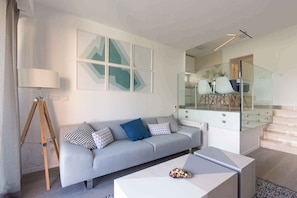 A modern living room with a grey sofa, a tripod floor lamp, and a coffee table. A staircase with glass railings leads to an upper level.