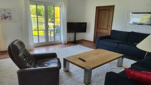 Un salon avec un fauteuil en cuir, un canapé bleu, une table basse en bois, un téléviseur et une porte-fenêtre coulissante donnant sur un jardin.