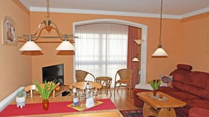 Una sala de estar con un sofá rojo, una mesa de comedor de madera y sillas. Hay un televisor, una araña y una ventana con cortinas.
