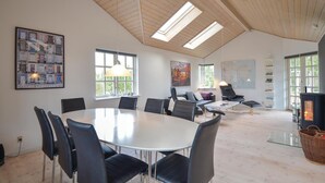 Een ruime kamer met een hoge plafond, een ronde tafel met stoelen, een open haard en grote ramen.