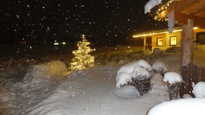 A snow-covered landscape with a lit Christmas tree and a building with a warm glow.