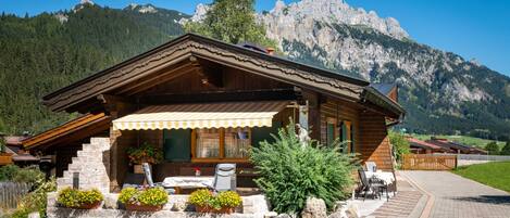 A wooden chalet with a stone foundation, a covered porch, outdoor seating, and a mountain backdrop.